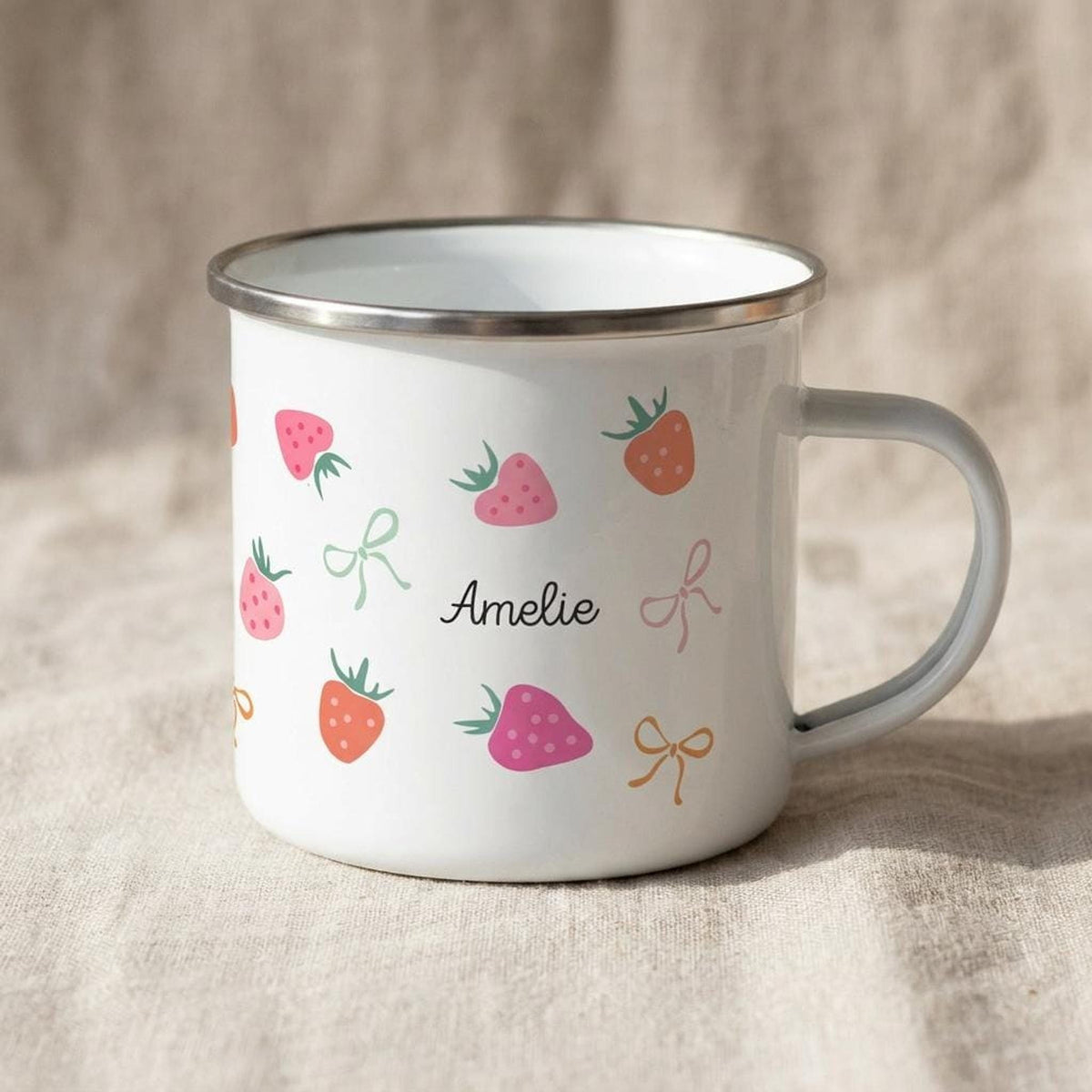 A white enamel mug with a metallic rim, adorned with strawberry graphics and the name Amelie printed in black script, set on a draped linen background.