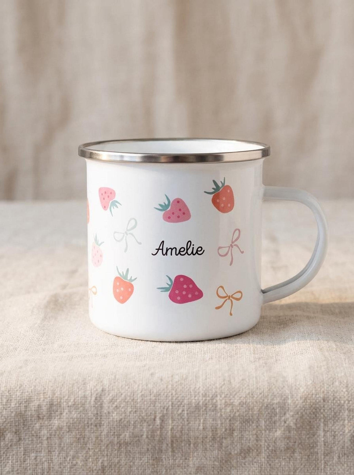 A white enamel mug with a silver rim, featuring strawberry motifs, bows, and the custom name Amelie centered on the side.