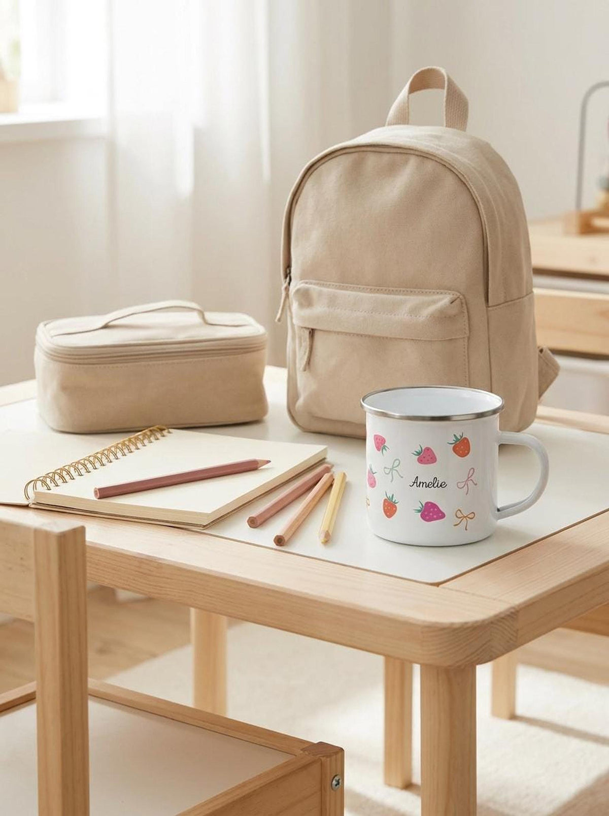 A child's wooden desk displays a personalised white enamel mug with strawberries, a spiral notebook, colored pencils, a pouch, and a small backpack.