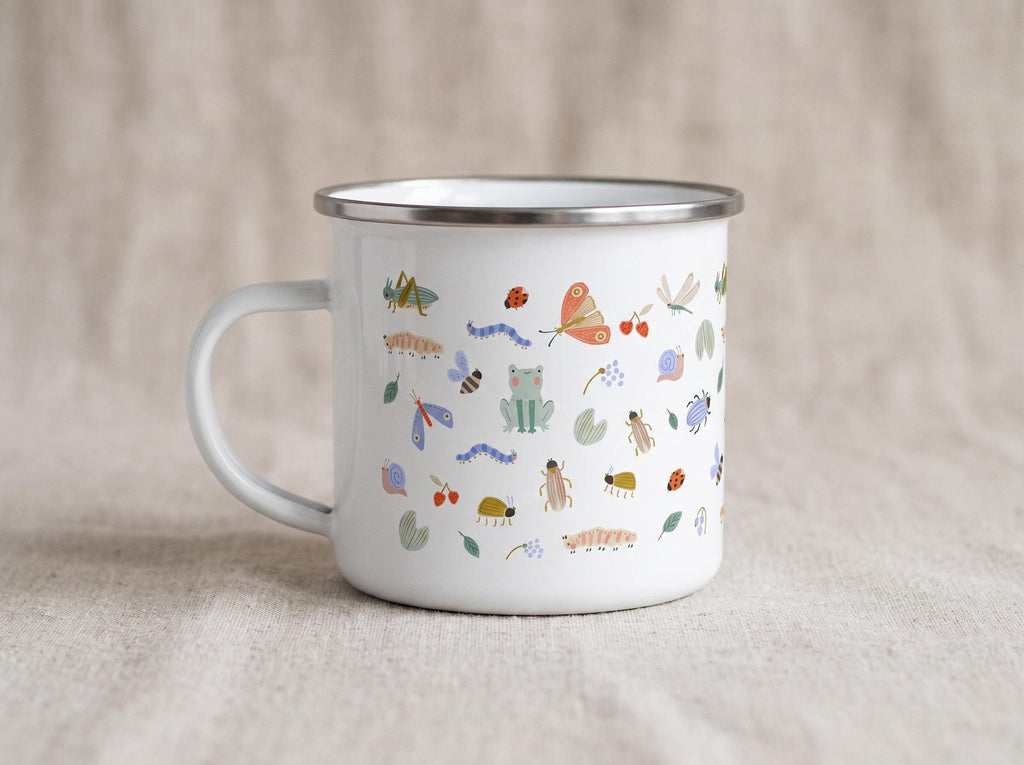 White enamel mug featuring an all-over pattern of colorful garden bugs and leaves, shown on a neutral fabric background.