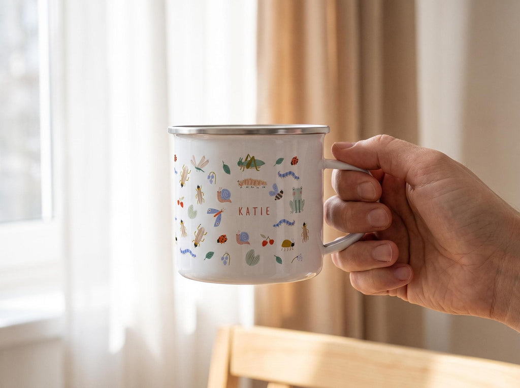 Hand holding a white custom enamel mug with a bug pattern and the name KATIE, against a window and curtain background.