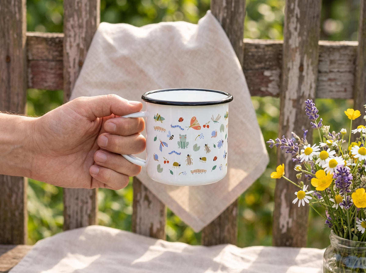 Hand holding a white enamel mug with bug illustrations in front of a wooden fence and a jar of wildflowers.
