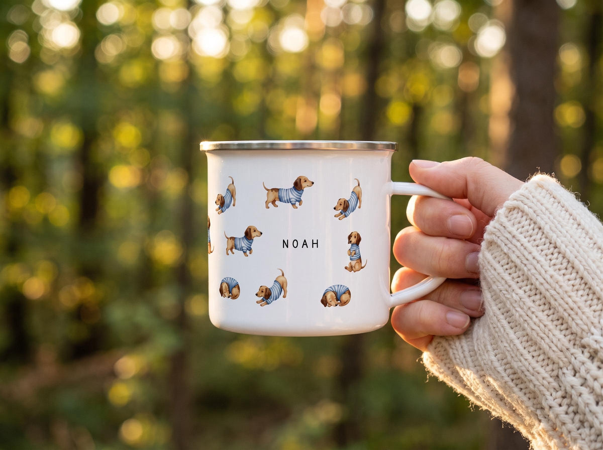 A hand wearing a cream knit sweater holds a white enamel mug decorated with illustrations of dachshunds in blue striped sweaters and the name Noah.