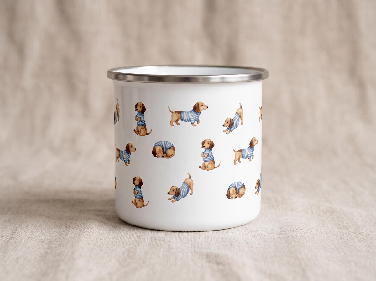 A white enamel mug with a silver rim displays a scattered pattern of illustrations of dachshunds in striped shirts against a neutral textile background.