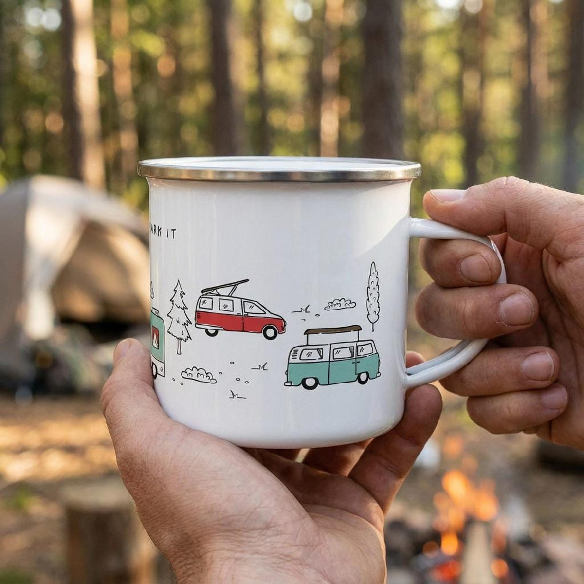 Hands holding the white enamel campervan mug outdoors with tents and trees in the background.