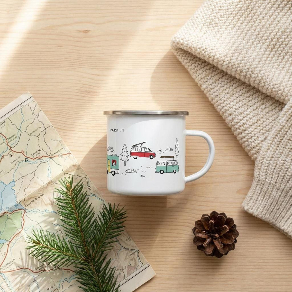 Flat lay showing the campervan enamel mug on a wooden table next to a map, pine branch, pinecone, and folded knit fabric.