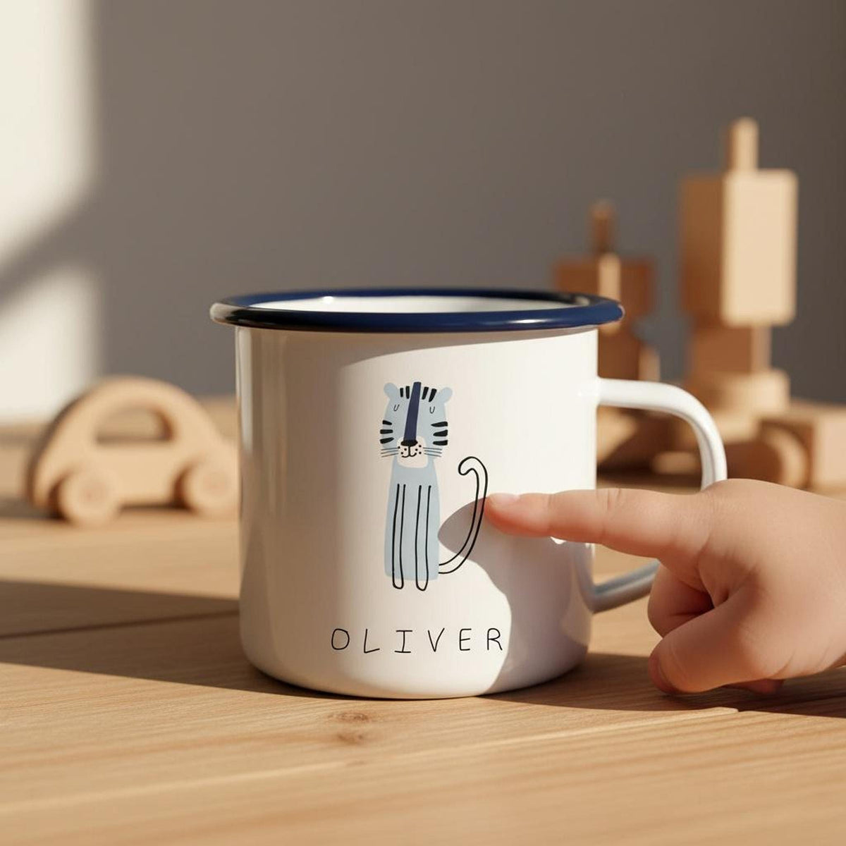 A child's hand points to a blue tiger illustration on a white enamel mug with a dark blue rim on a wooden surface.