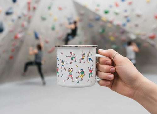 A hand holds a white enamel mug decorated with colorful rock climbers against an indoor climbing gym background.