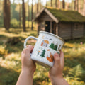 A child's hands hold a white enamel mug with colorful illustrations of trees, a house, and an orange fox, outdoors with a blurred log cabin in the background.