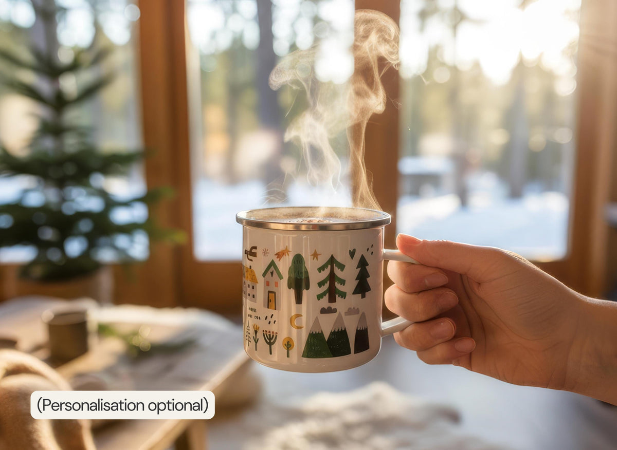 A hand holds a steaming white enamel mug printed with trees, a house, and flowers, in front of a bright window with a snowy view.
