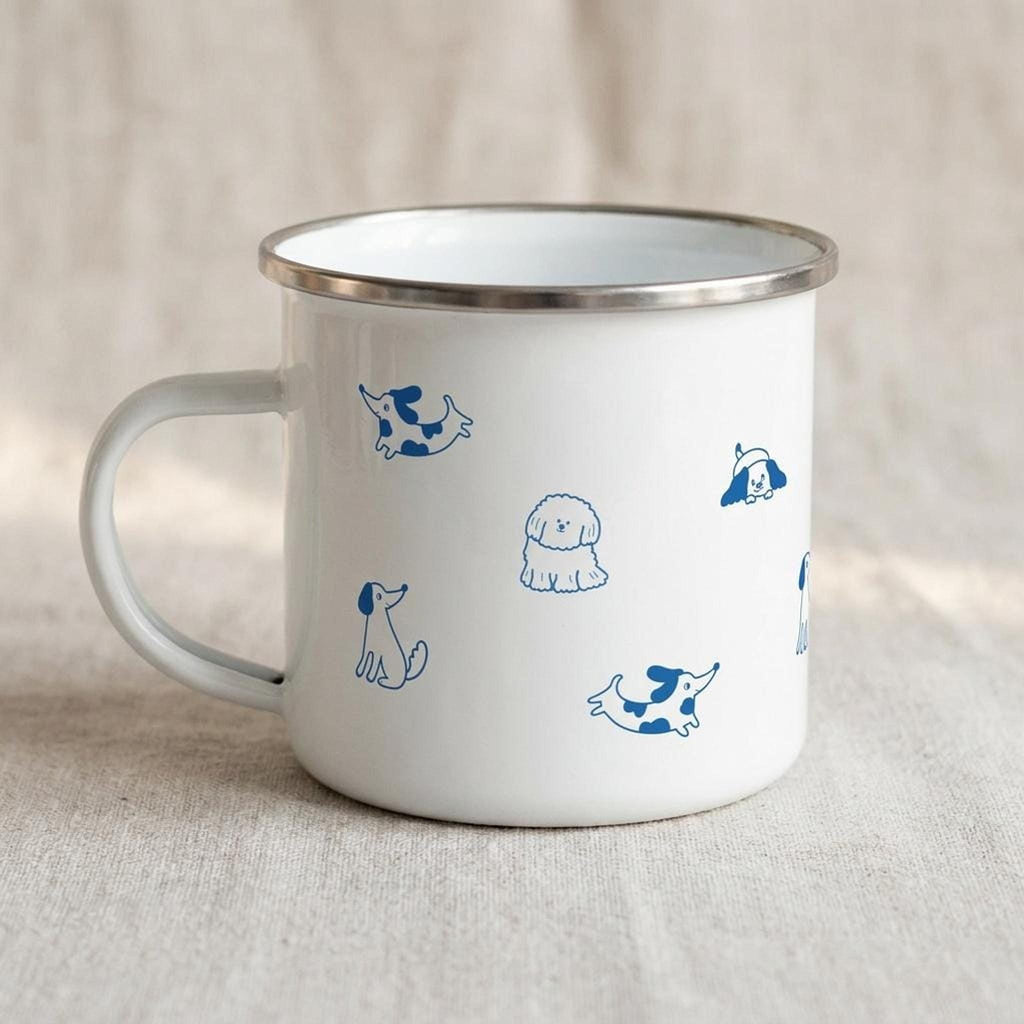 A white enamel mug with a silver rim and blue illustrated dog patterns sits on a neutral fabric surface, viewed from the handle side.