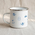 A white enamel mug with a silver rim and blue illustrated dog patterns sits on a neutral fabric surface, viewed from the handle side.