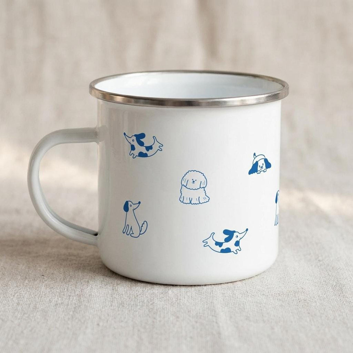 A white enamel mug with a silver rim and blue illustrated dog patterns sits on a neutral fabric surface, viewed from the handle side.