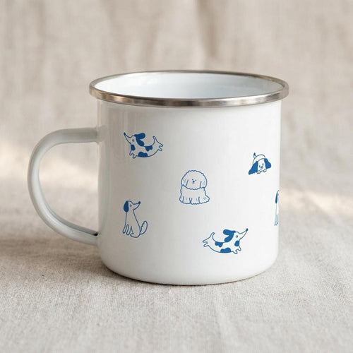 A white enamel mug with a silver rim and blue illustrated dog patterns sits on a neutral fabric surface, viewed from the handle side.