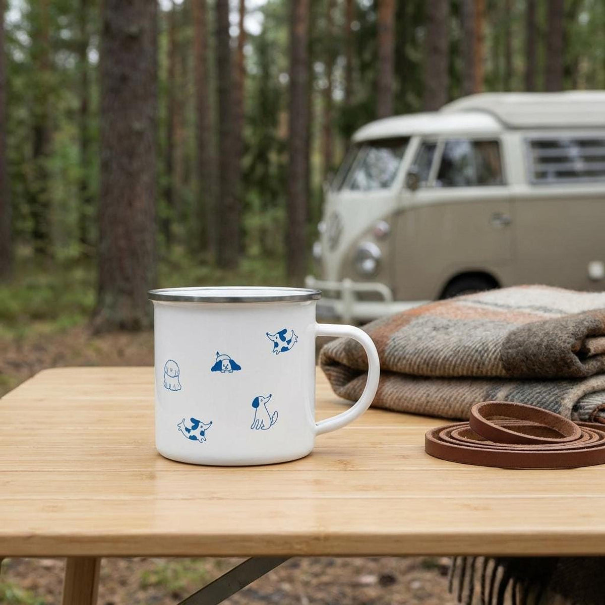 A white enamel mug with blue dog illustrations sits on a wooden camping table outdoors, with a parked van and pine trees in the blurred background.