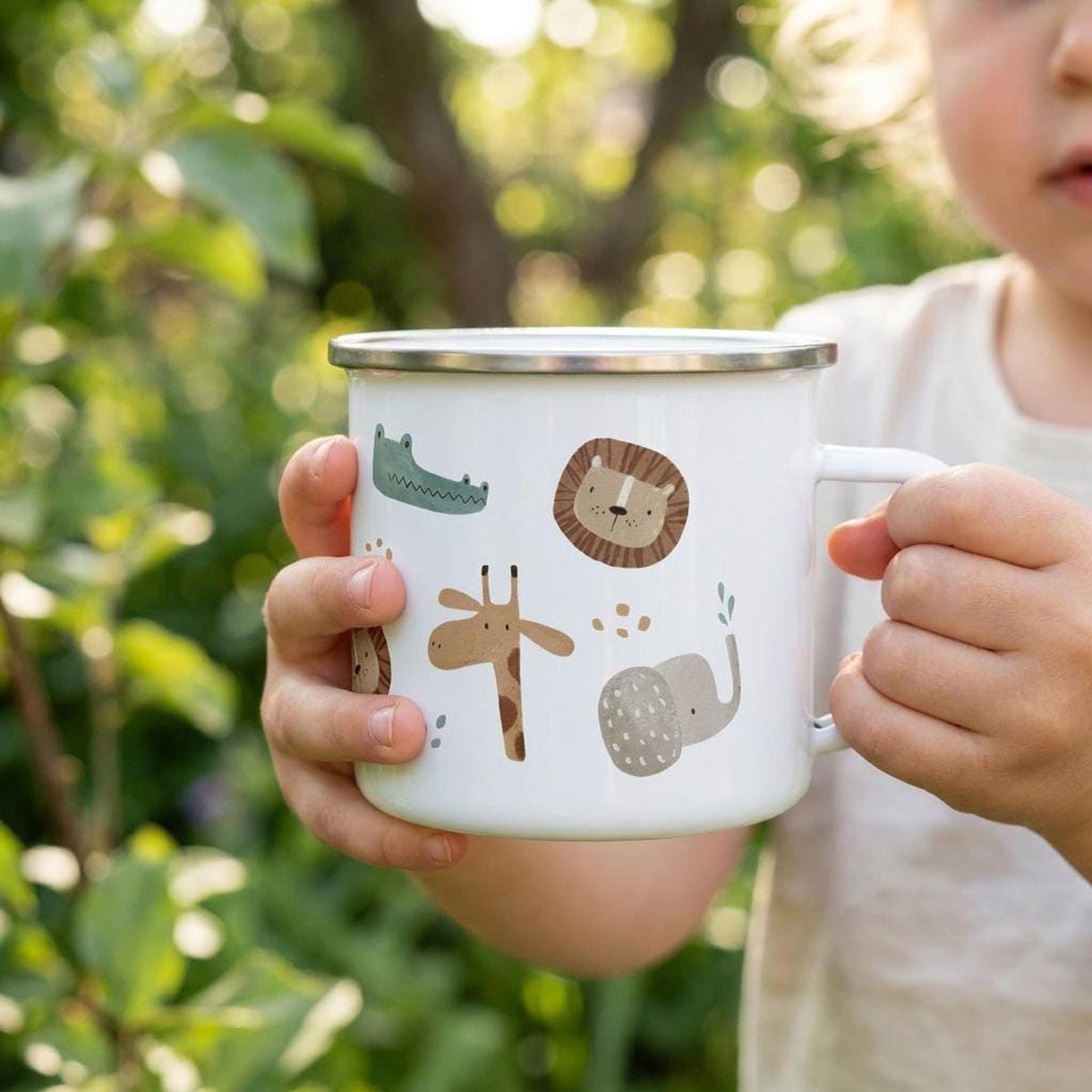 A child holds a white enamel mug decorated with illustrations of safari animals including a giraffe, lion, and elephant outdoors.