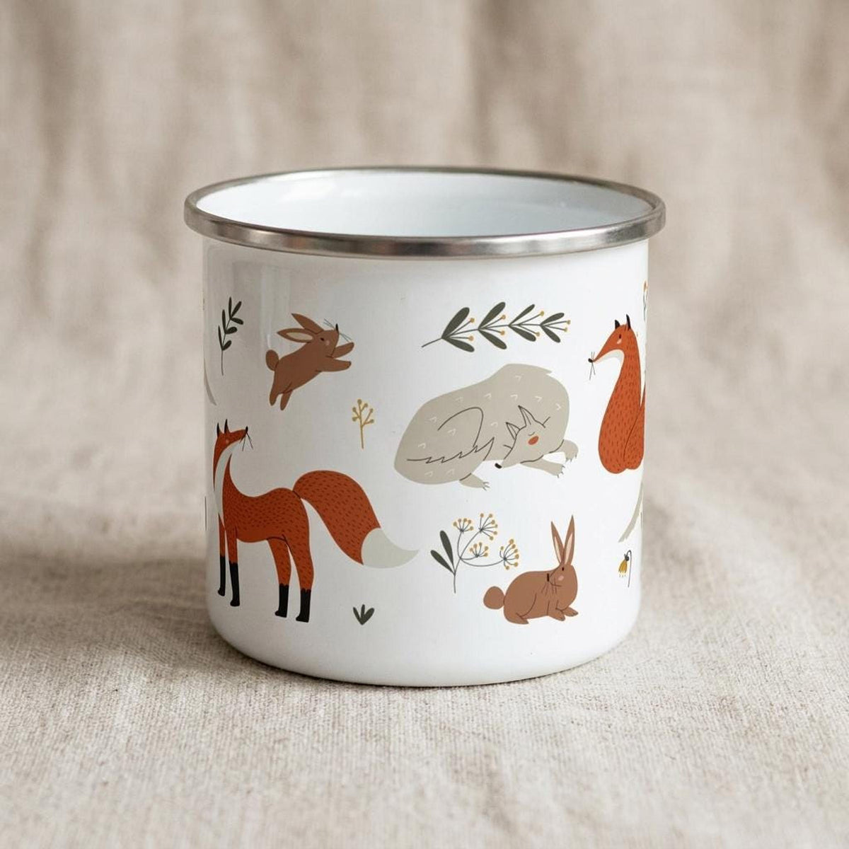 A white enamel mug with a silver rim shows a different angle of the forest animal pattern with a resting wolf, a standing fox, rabbits, and botanical elements.