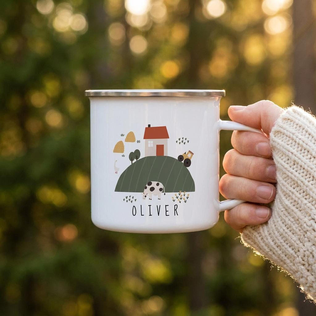 A hand in a knit beige sweater holds a white enamel mug featuring a farm scene and the name Oliver against a soft green out-of-focus background.