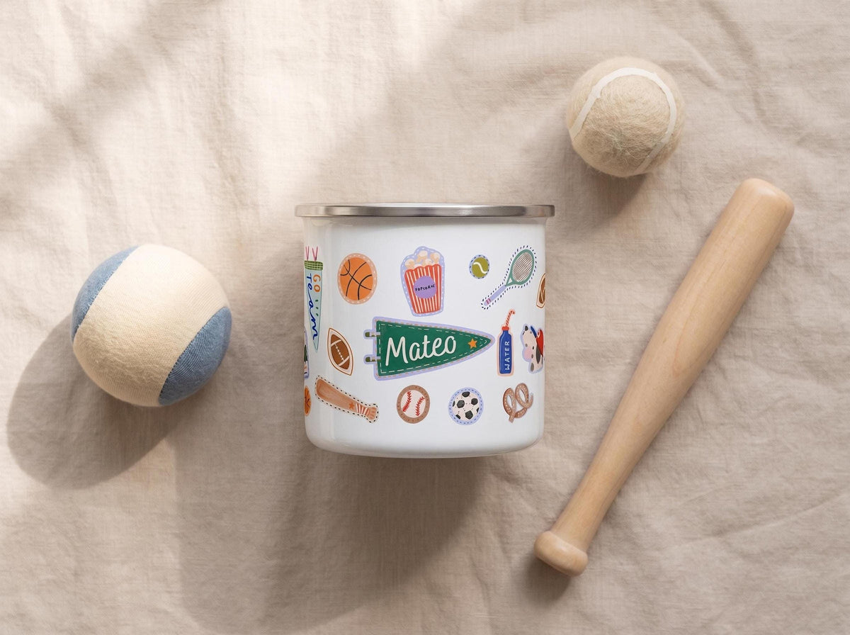 A white enamel mug with sporty stickers and the name Mateo printed on it, surrounded by toy sports balls and a mini wooden bat on a beige linen surface.