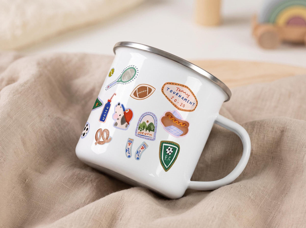A tilted white enamel mug resting on a crumpled beige cloth, displaying assorted sports stickers on its side.