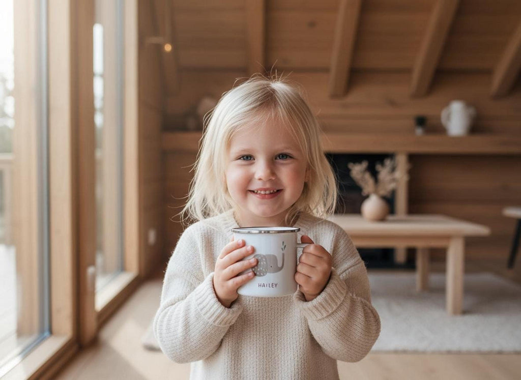 A young blonde child wearing a cream sweater smiling while holding a white enamel mug with an elephant graphic and the name Hailey.