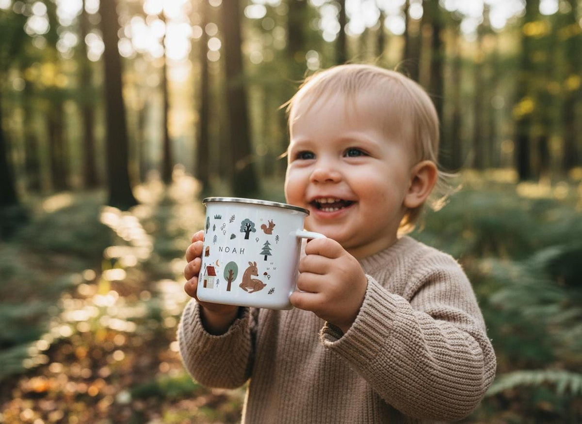 A smiling toddler holding a personalised woodland enamel mug with the name Noah in a sunlit forest.