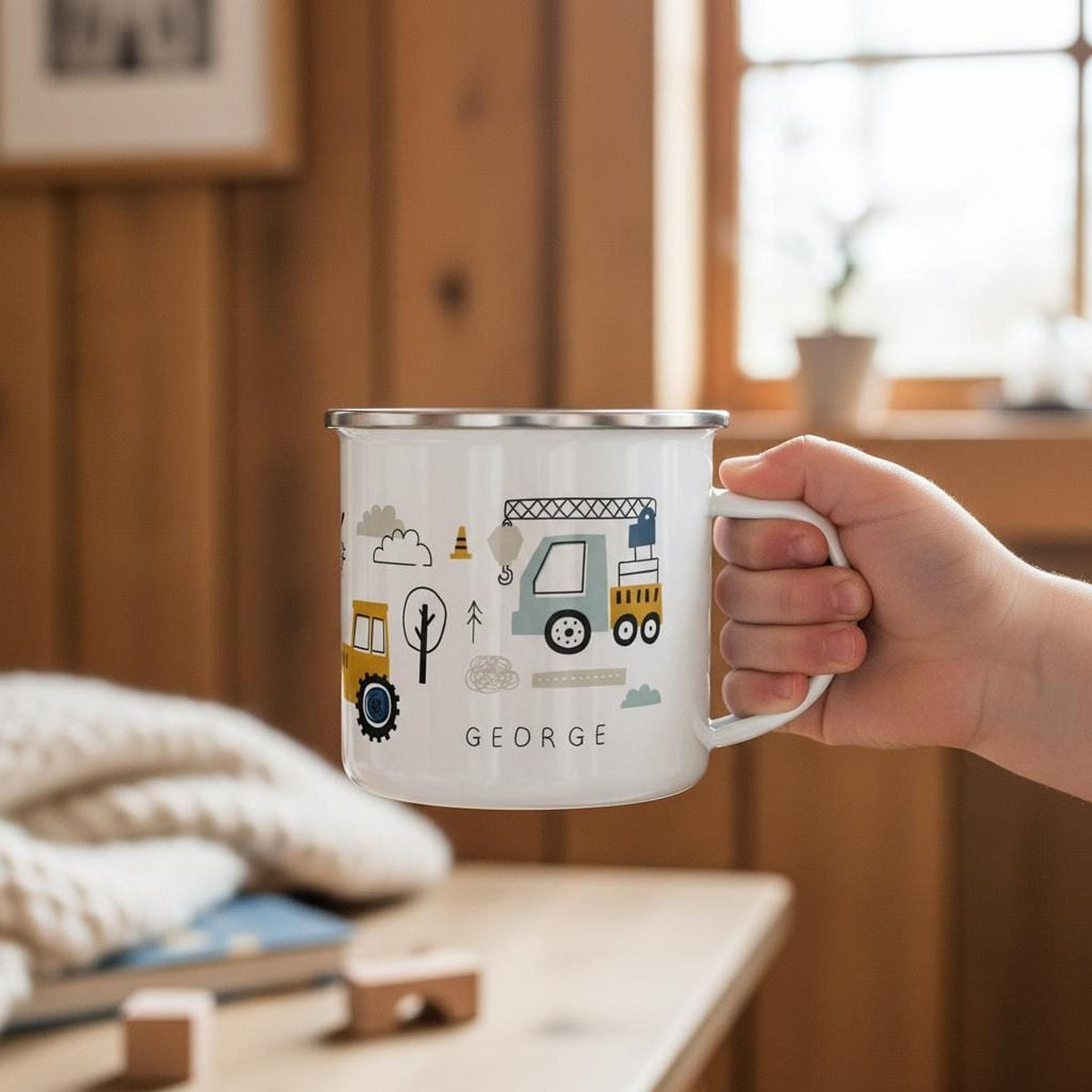 A hand holds a white enamel mug featuring a construction vehicle illustration and the name George, placed on a wooden table with a folded knit blanket in the background.