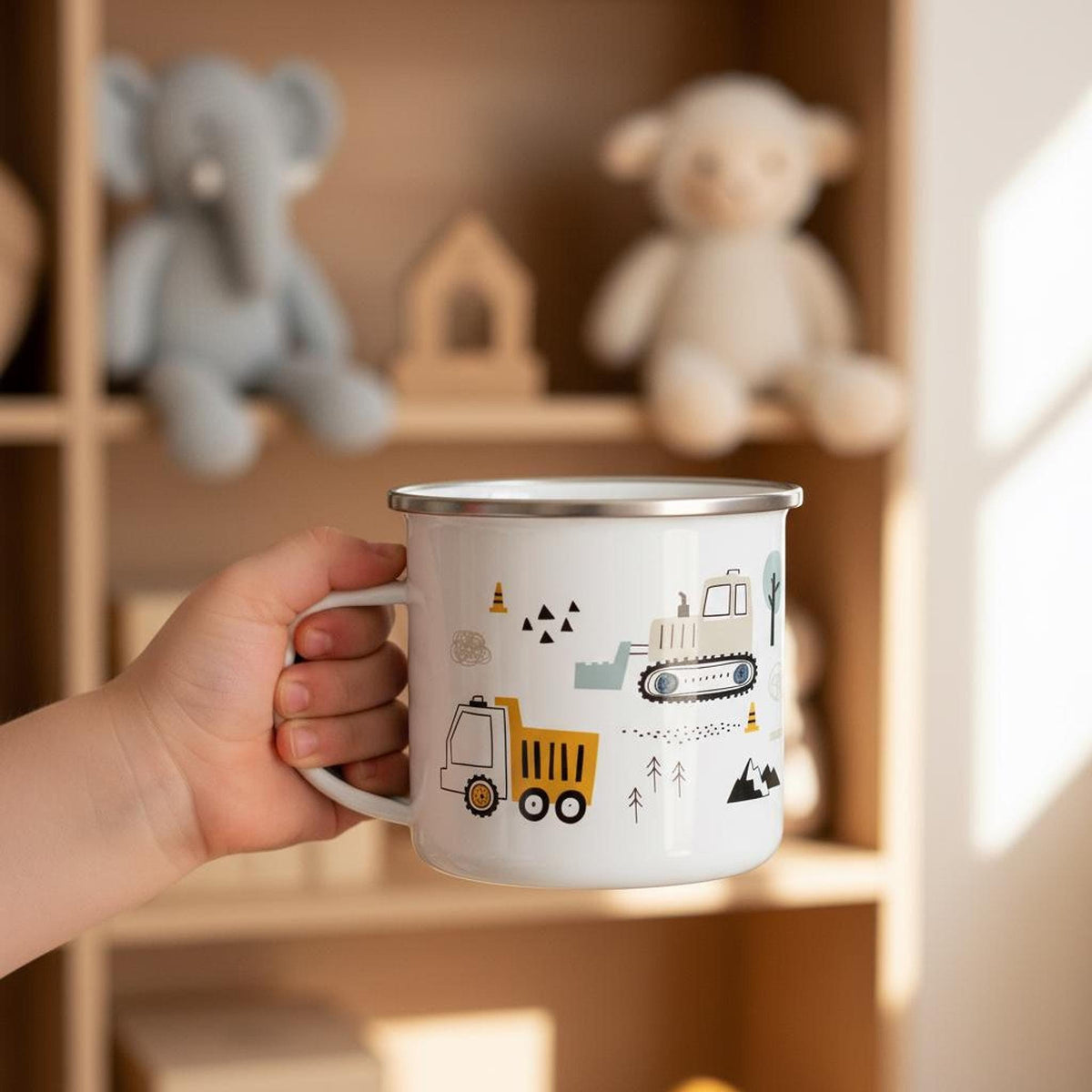 A child's hand holds a white enamel mug featuring a dump truck illustration in front of a wooden shelf holding stuffed animals and wooden toys.