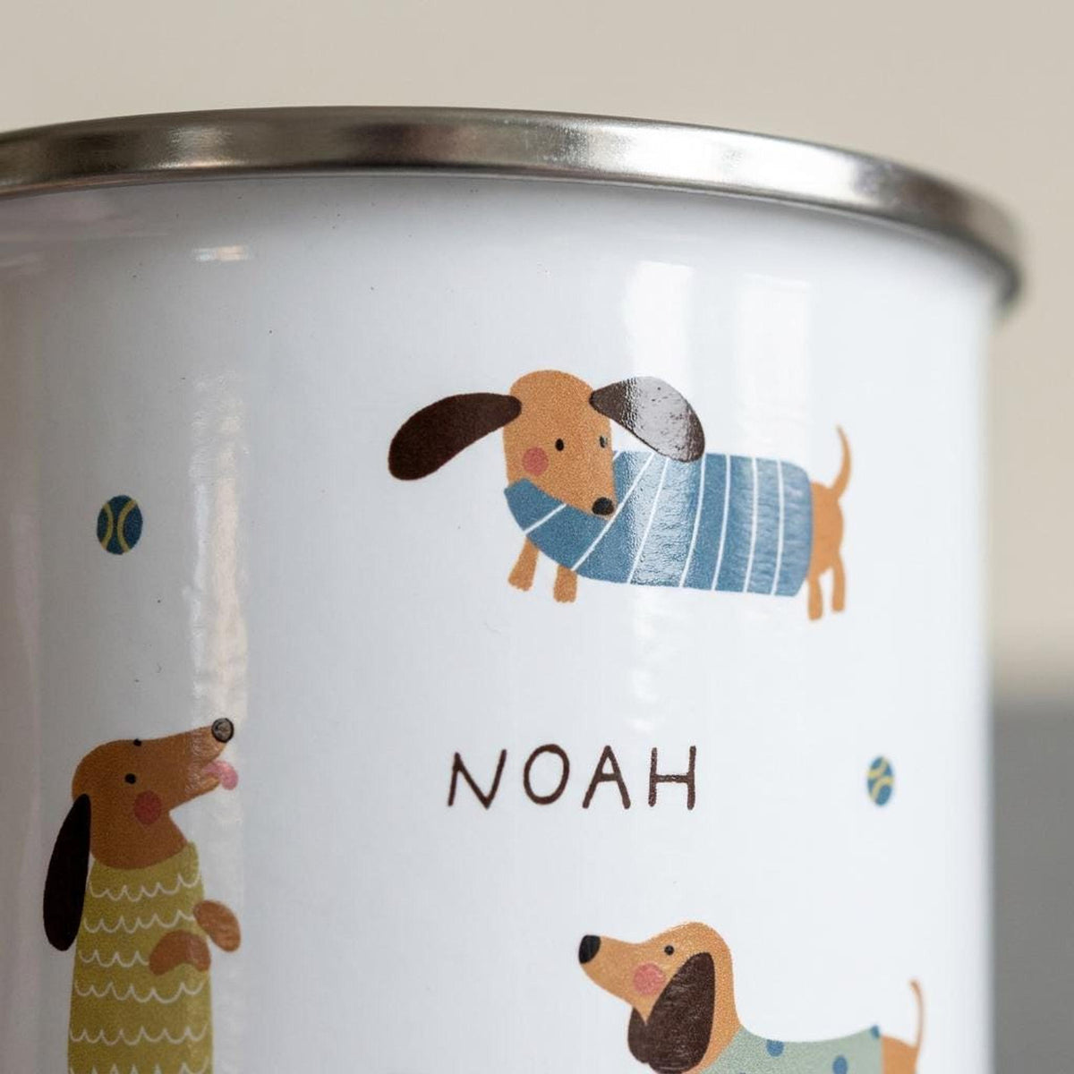 A close-up shot of the white enamel mug detailing one dachshund illustration and the name Noah printed on the side.