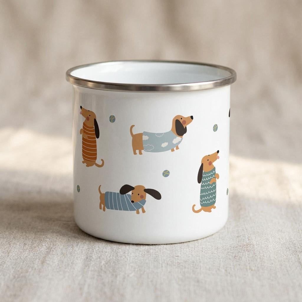 A white enamel mug printed with cartoon dachshunds in sweaters on a textured fabric backdrop.