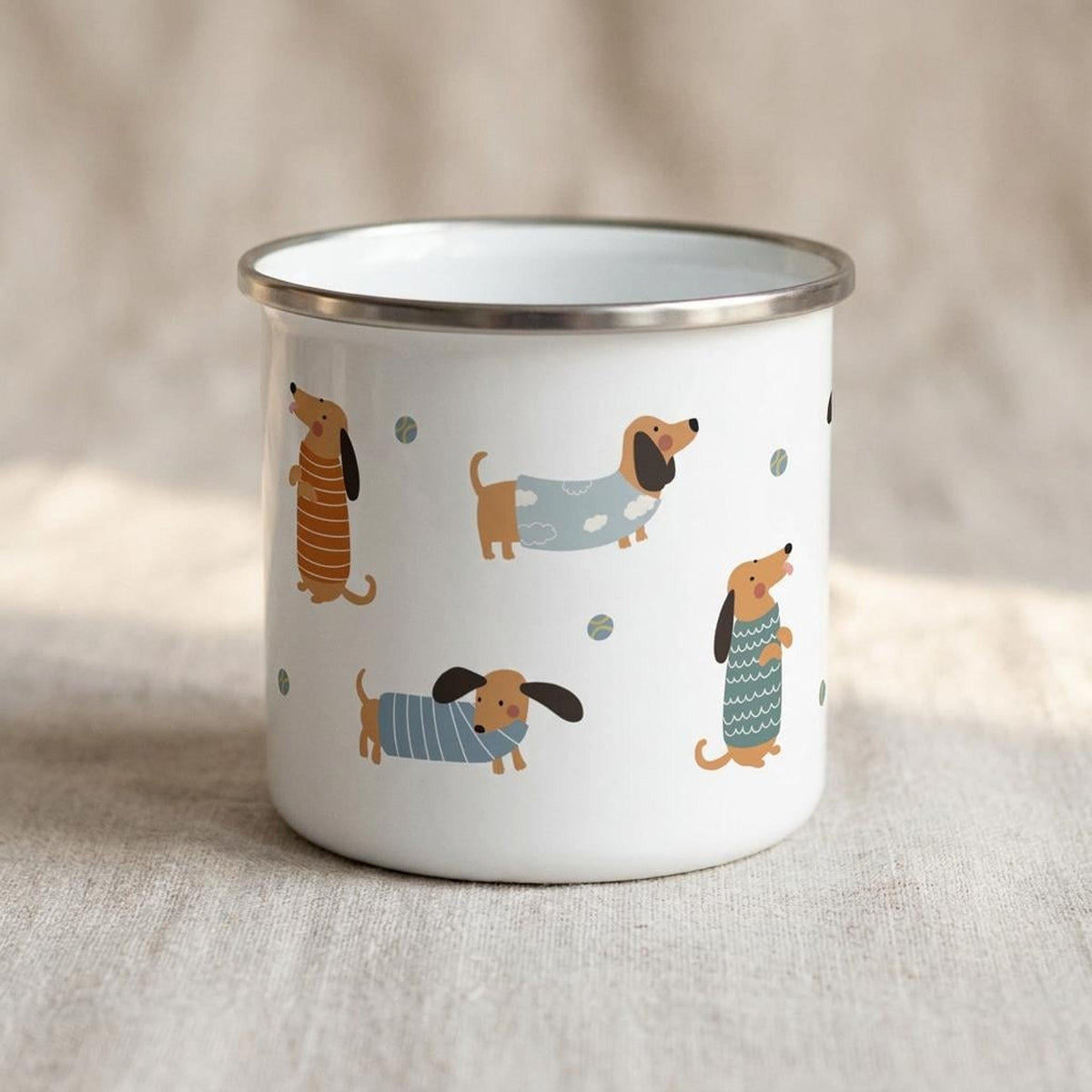 A white enamel mug printed with cartoon dachshunds in sweaters on a textured fabric backdrop.