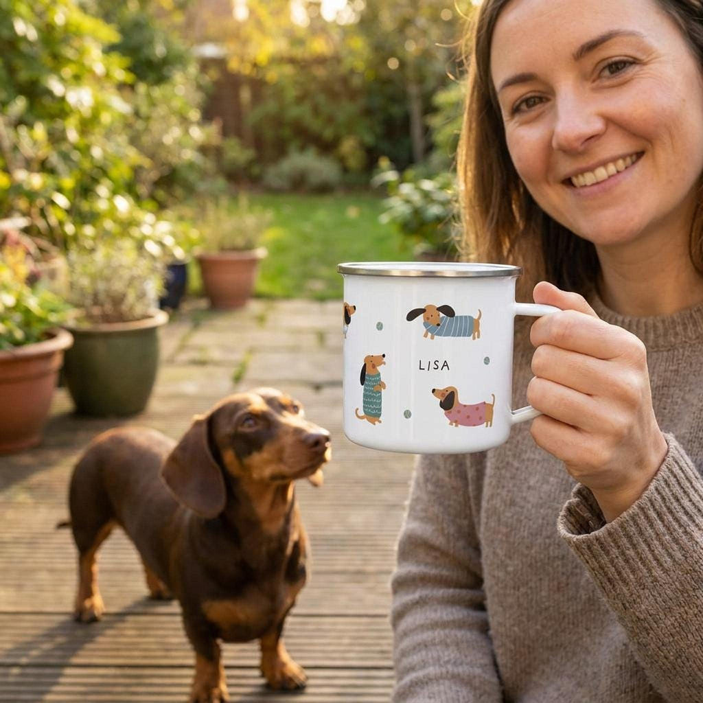 A smiling woman holds a personalized dachshund enamel mug outdoors on a deck while a smooth-haired dachshund looks up at her.