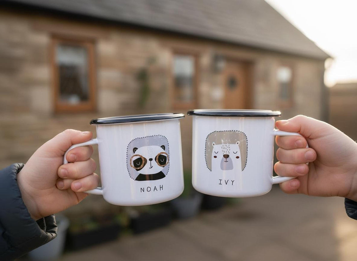 Two hands hold matching white enamel mugs with animal graphics and names in front of a stone building.