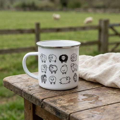 A white enamel mug with a silver rim and black cartoon sheep illustrations rests on a rustic wooden table with a blurred green field and sheep in the background.