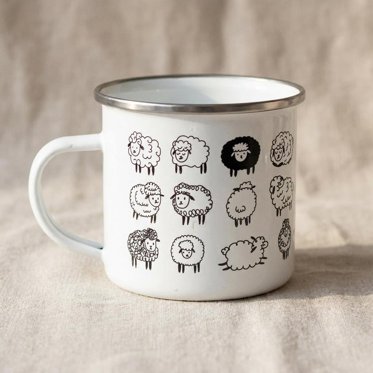 A white metal enamel mug with a silver rim displays a grid pattern of cute black cartoon sheep illustrations against a neutral textured fabric background.