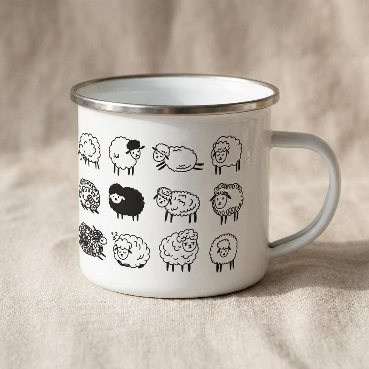 A white enamel mug with a silver rim and handle is printed with multiple black sheep illustrations, set against a light brown textured backdrop.