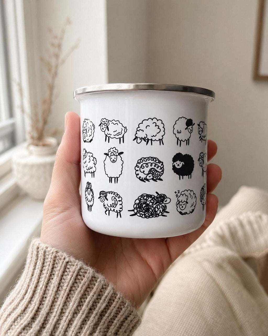 A person holds a white enamel mug printed with a pattern of black sheep against a bright window and neutral background.
