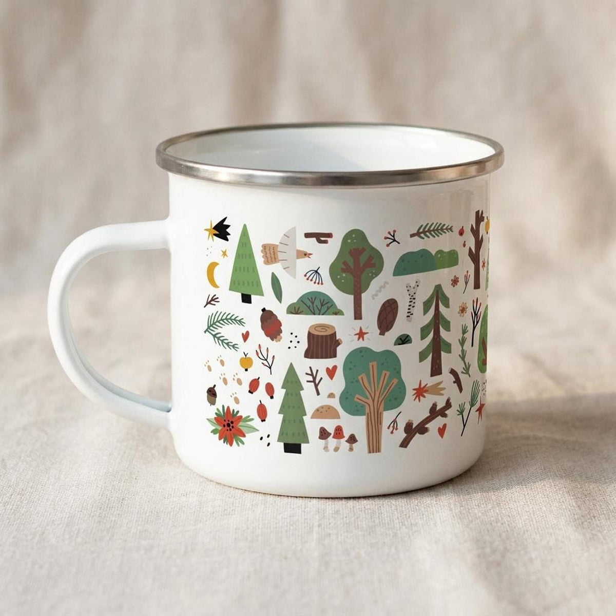 A white enamel mug with a silver rim sits on a beige fabric surface, displaying an illustrated forest pattern including trees, birds, and pinecones.