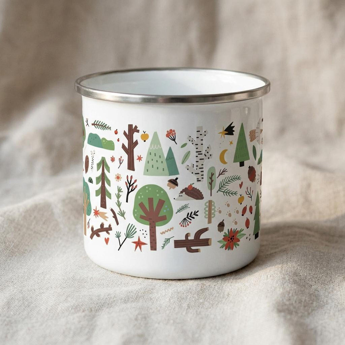 A white enamel camping mug featuring an illustrated woodland pattern stands on a neutral textured fabric.