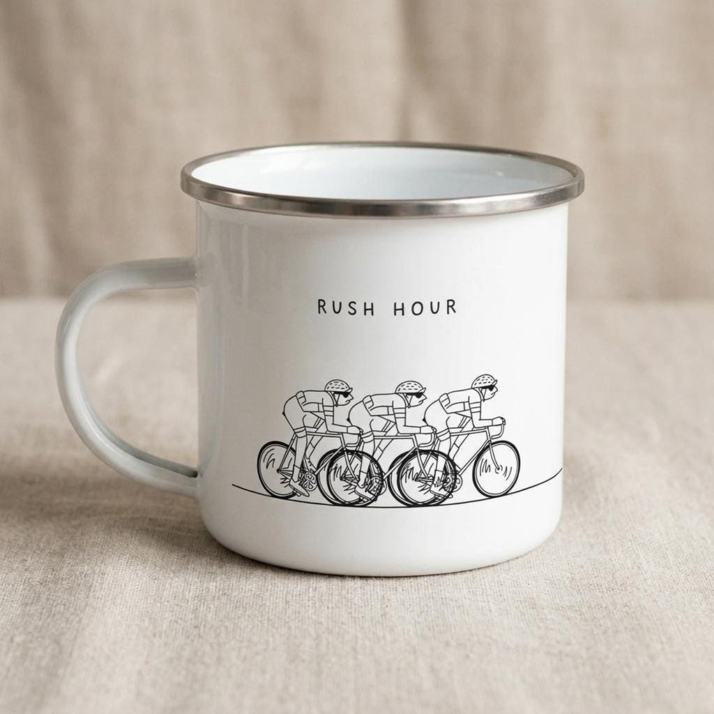 A white enamel mug with a silver rim displays a black illustration of four cyclists and the text RUSH HOUR.