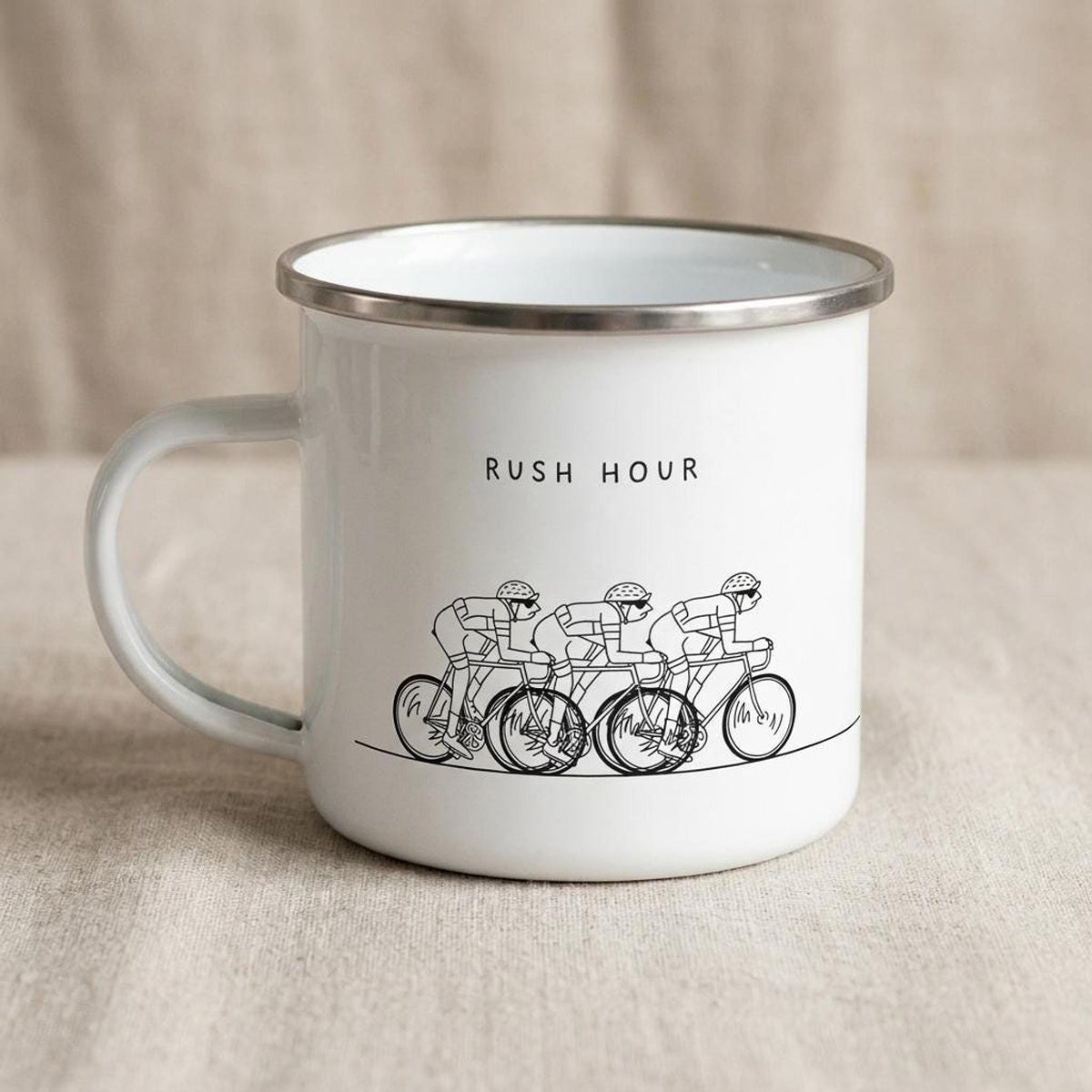 A white enamel mug with a silver rim displays a black illustration of four cyclists and the text RUSH HOUR.
