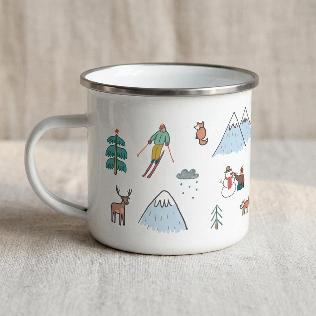 A white enamel camp mug with a silver rim displays colorful illustrations of mountains, trees, a skier, animals, and a snowman, sitting on a textured beige fabric surface.