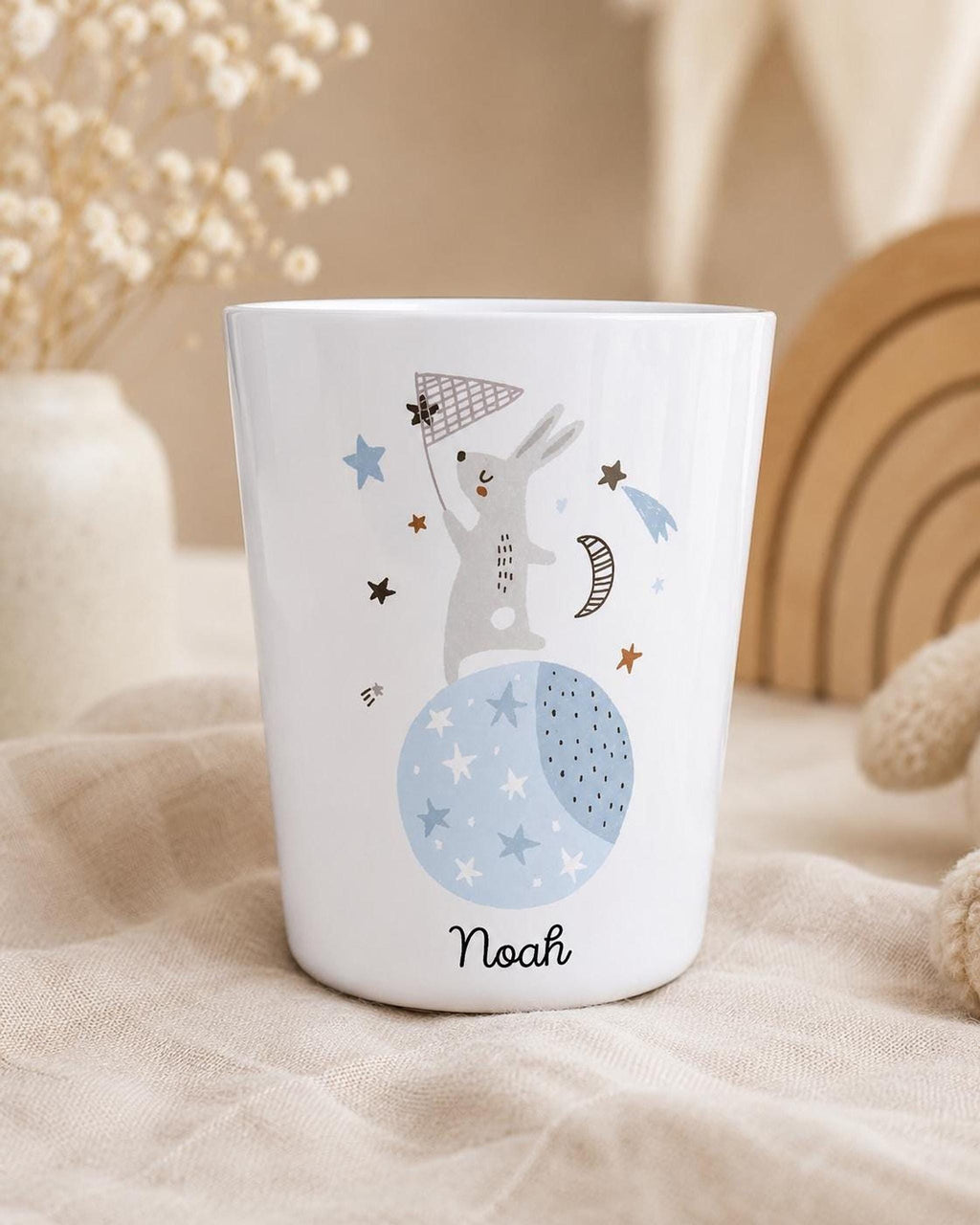 A close-up of the white cup with the bunny and moon graphic and the name Noah, set on a fabric background with a vase of dried flowers and a wooden rainbow in the background.