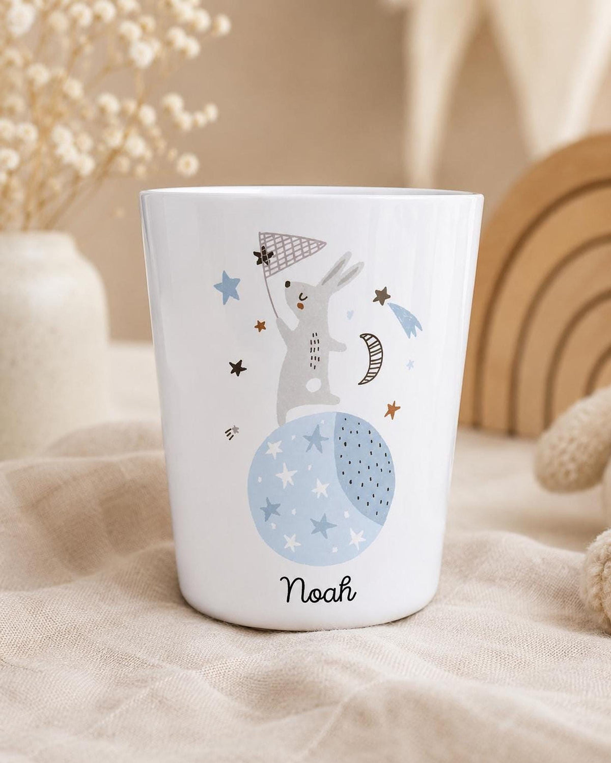 A close-up of the white cup with the bunny and moon graphic and the name Noah, set on a fabric background with a vase of dried flowers and a wooden rainbow in the background.