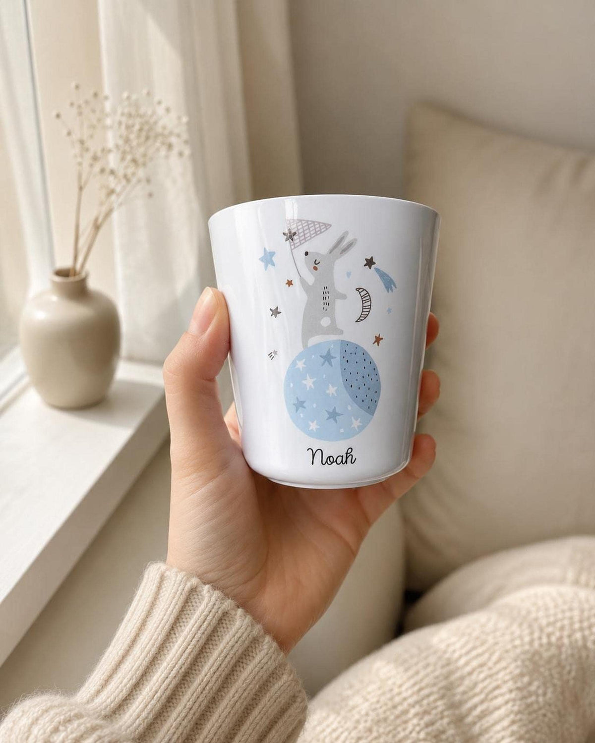 A hand wearing a cream knit sweater holds the white personalised bunny cup near a window with a vase of dried flowers and a pillow.