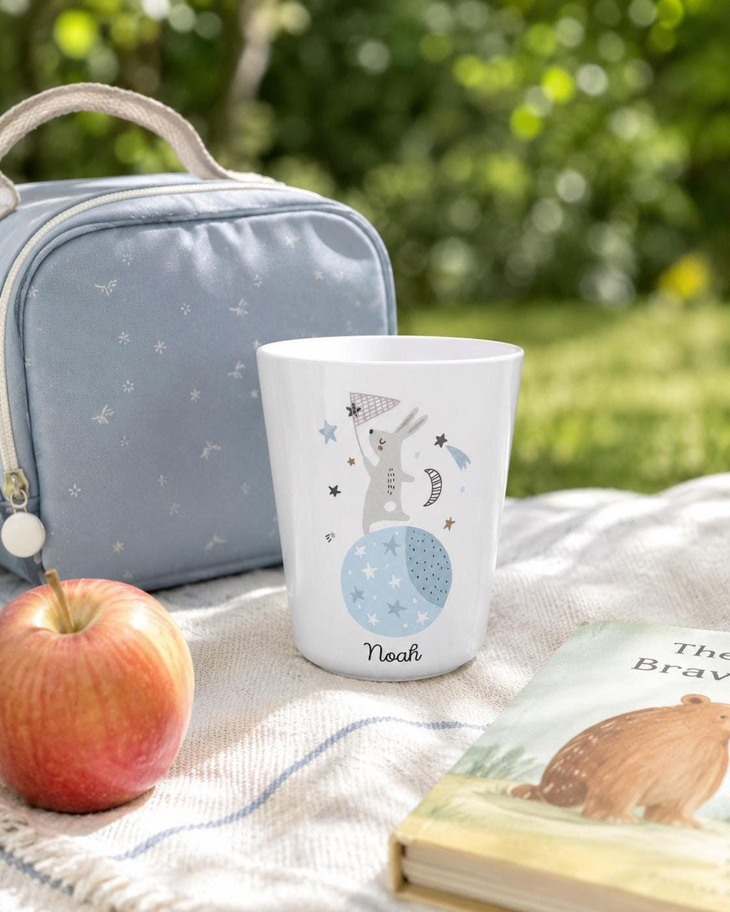 An outdoor picnic scene featuring the white personalized bunny cup sitting on a blanket next to a red apple, a blue bag, and a children's book.