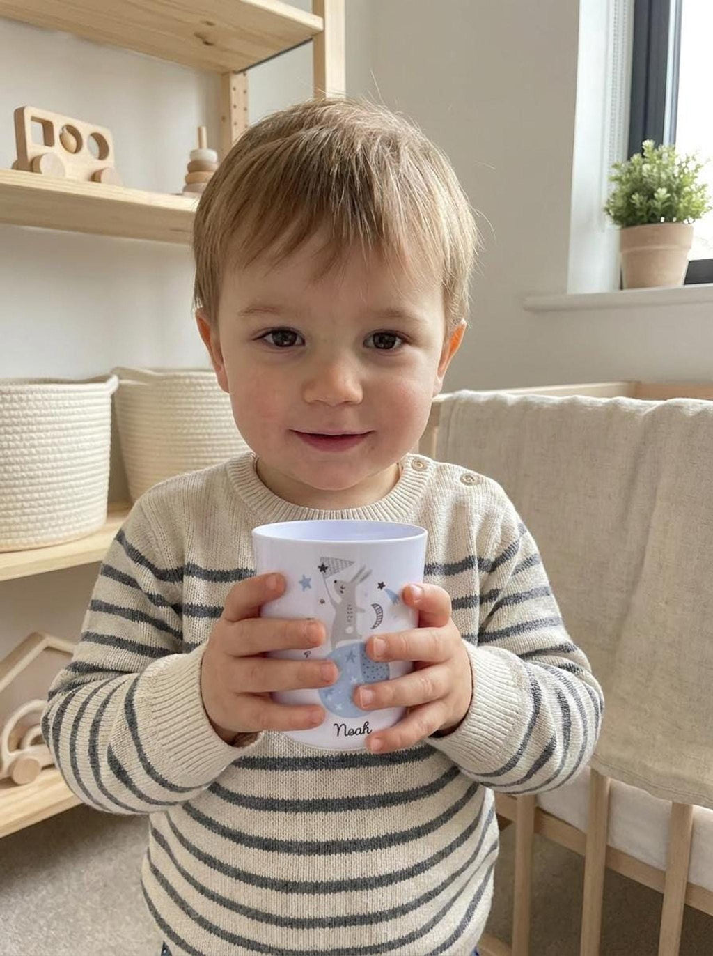 A young toddler with short blonde hair wearing a striped sweater holds the personalized white bunny cup in a nursery setting with a crib and storage baskets.