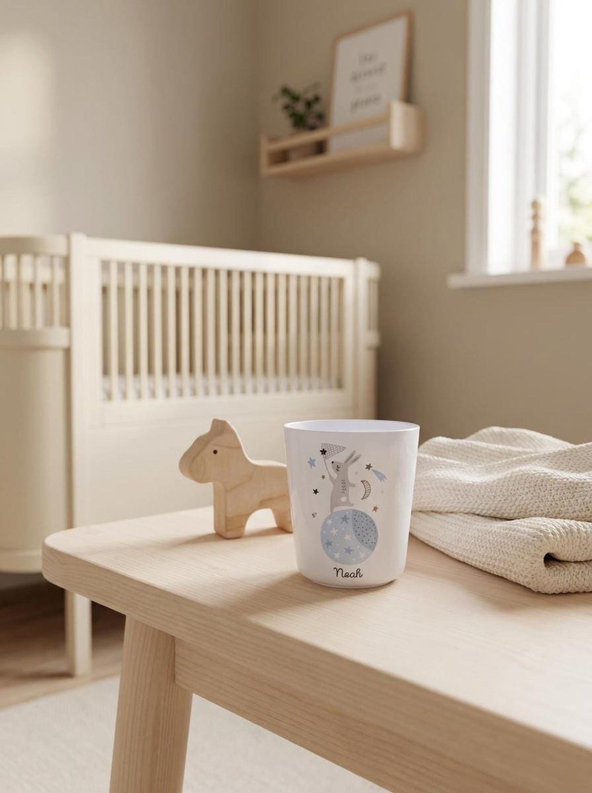 A wooden table in a nursery features the personalized white bunny cup alongside a small wooden toy horse and folded textile, with a crib in the background.