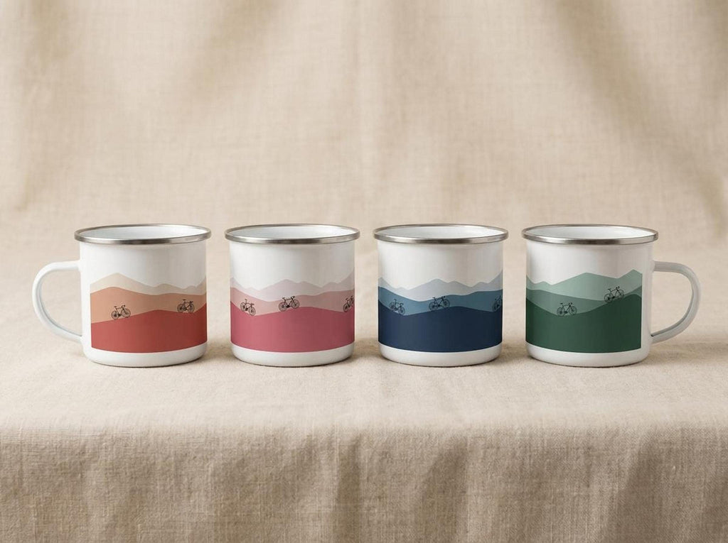 Four white enamel mugs featuring different landscape colorways lined up on a neutral fabric surface.