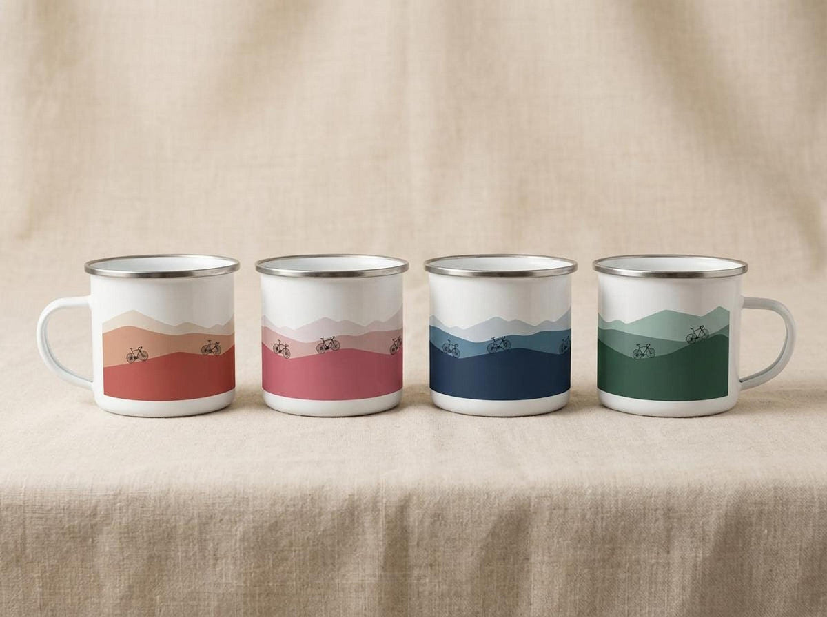 Four white enamel mugs featuring different landscape colorways lined up on a neutral fabric surface.